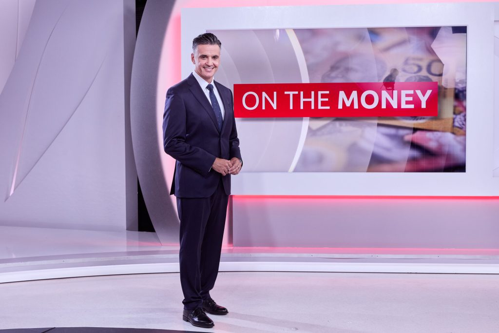 SBS Finance Editor Ricardo Gonçalves wearing a black suit standing infront of the SBS news desk that is white with red trim lighting and the words "On the money" in red text in the background.