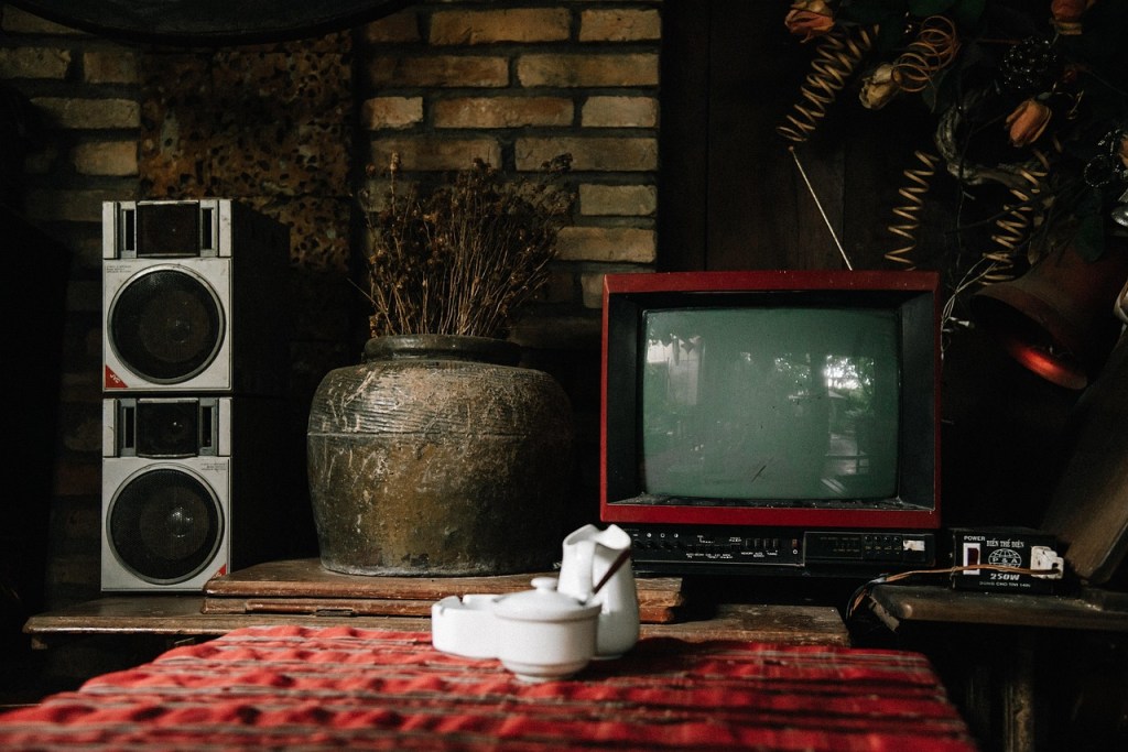 A dark living room of a switched off television and speakers to the left. In the foreground is a white teapot and mug on a red coffee table.