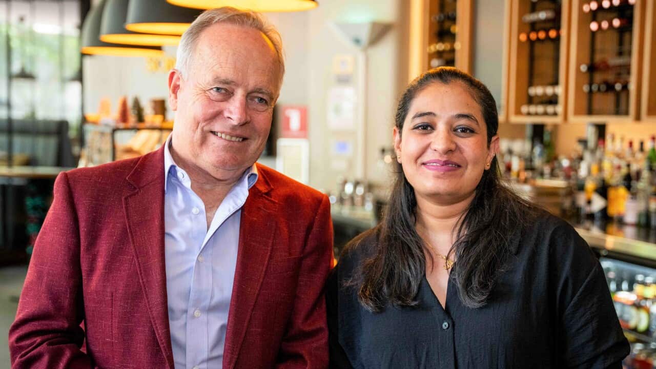 A man in a burgundy jacket stands in a bar next to a woman with long brown hair in a dark blue shirt. 