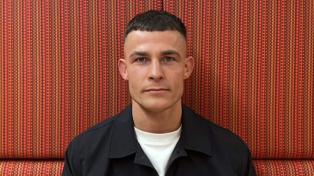 a closeup of a young man in a white tshirt and black jacket sits in front of a red patterned background