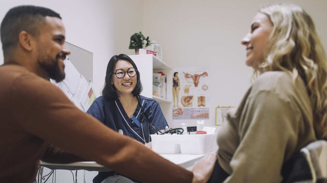 Happy female gynecologist looking at smiling man touching stomach of pregnant woman in clinic