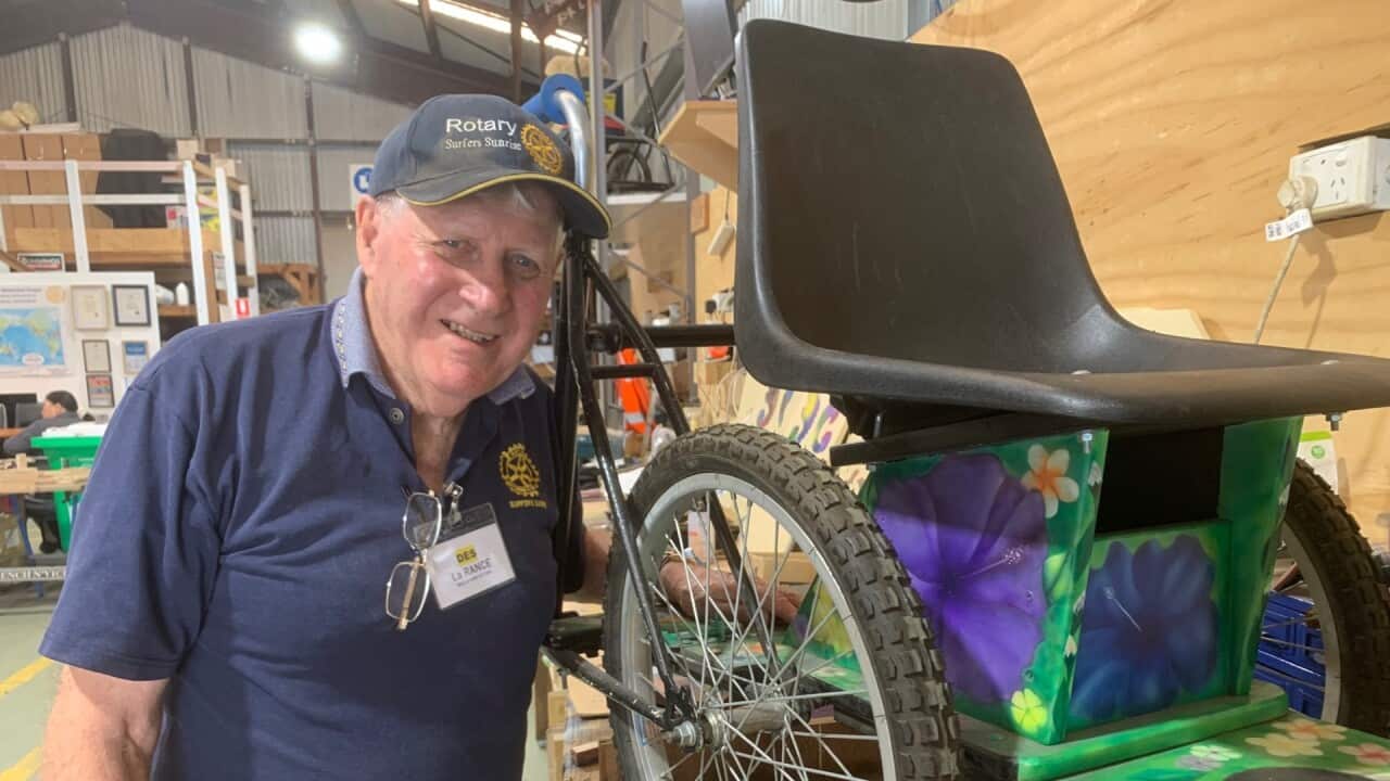 A man in a cap wearing a blue t-shirt stands next to a wheel chair made from bicycle parts.