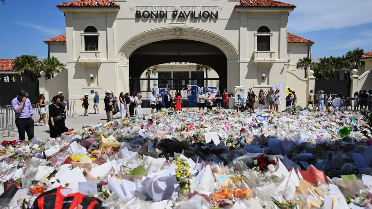 A few people stand around a huge amount of flowers laid in front of the gates of Bondi Pavilion.