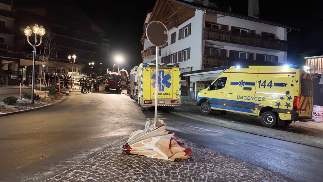 Emergency vehicles, including yellow ambulances and fire trucks, are stationed on a lit street at night in a snowy resort town following an incident.