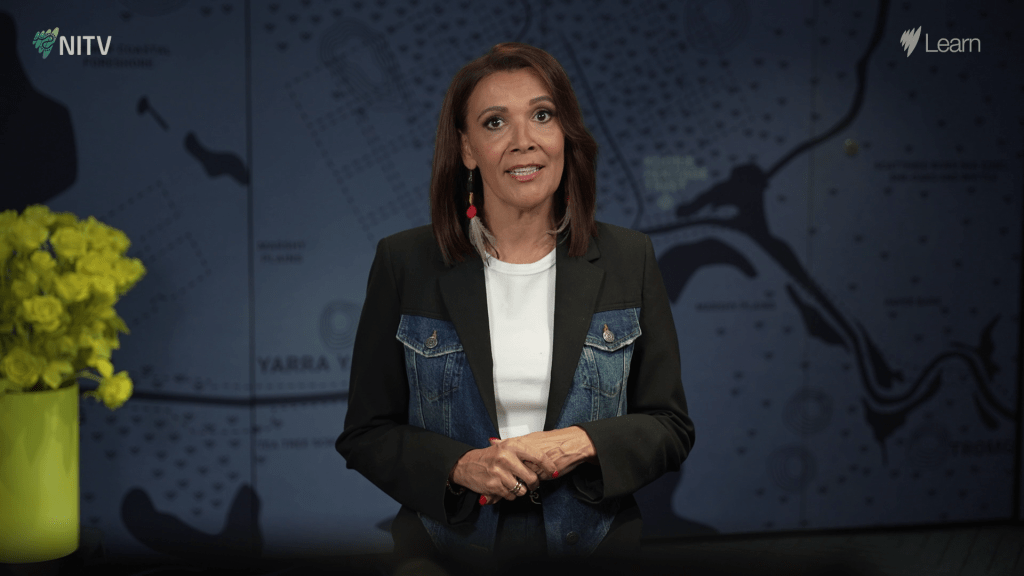 A First Nations educator woman, named Shelley Ware, wearing a black and denim blazer standing directly to camera with a blue backdrop of a First Nations outline map.